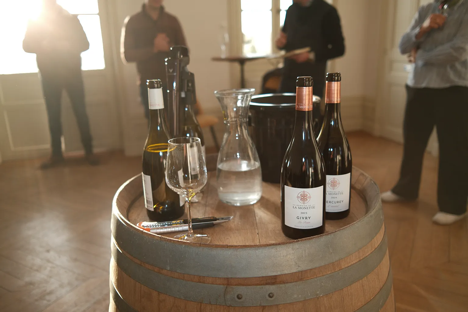 An image on winebottles in sunlight on a wine barrel indoors with people behind it.